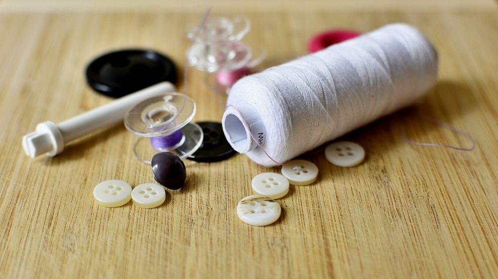 A spool of white thread, buttons, bobbins with colored thread, and a seam ripper are arranged on a wooden surface—perfect tools for quick fixes after a wardrobe malfunction.